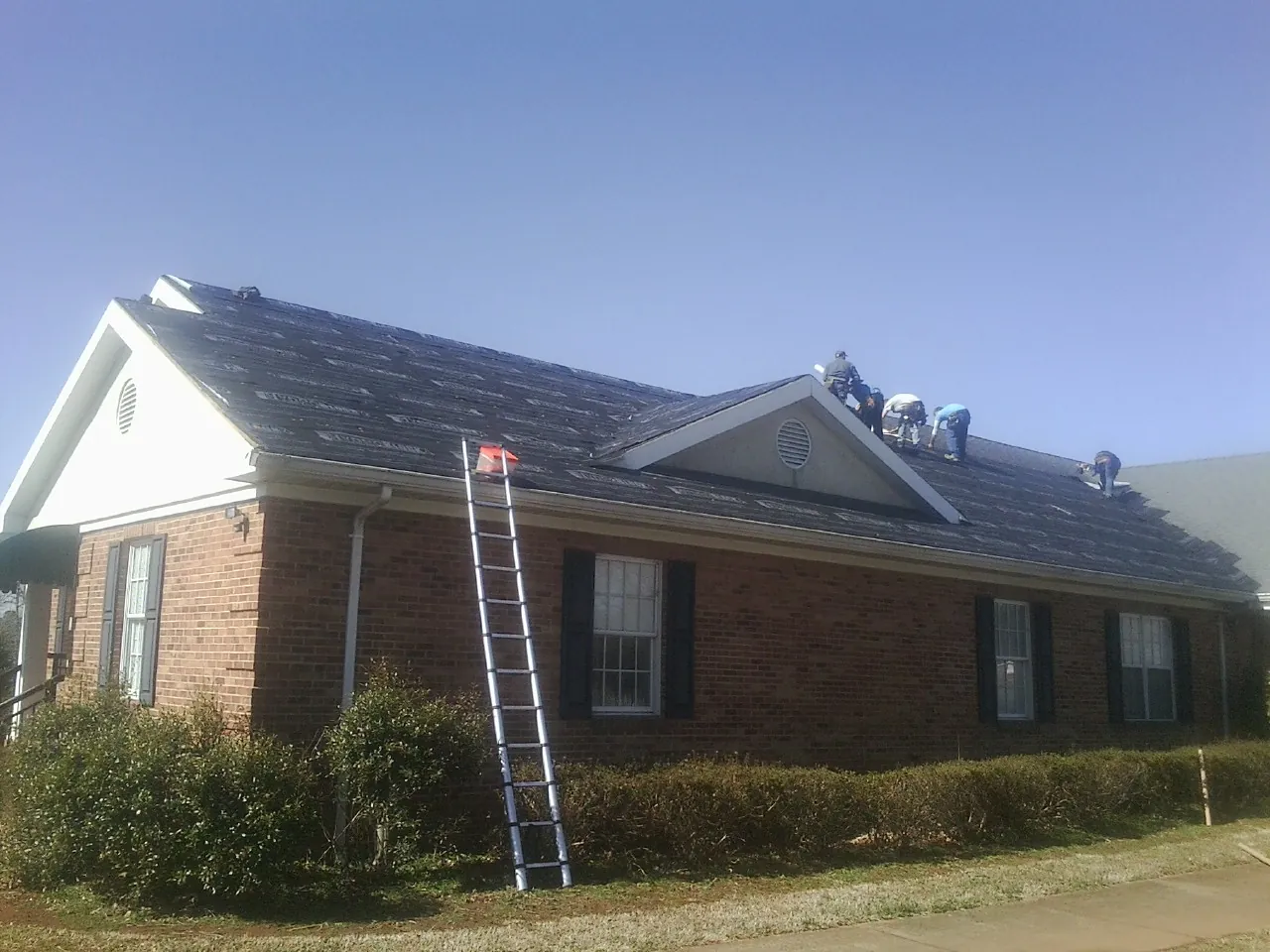 Crew tearing shingles off a brick ranch roof with a ladder set — Emergency Roof Repair work by our crew in Goldsmith