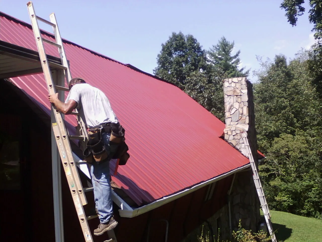 Roofer on a ladder beside a red metal roof and stone chimney — Soffit & Fascia Repair by Roofing Brothers in Goldsmith